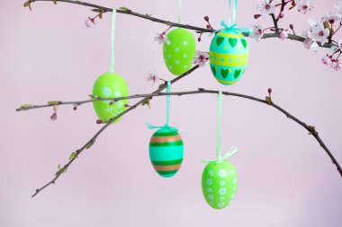 Kurdeleli yeşil ve sarı Paskalya yumurtaları çiçek açan bir ağaç dalından pastel arka planda sallanıyor. Bahar, kutlama ve yenilenmenin bayram ve dekoratif sembolü..