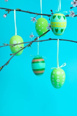 Çiçekli ve çizgili yeşil Paskalya yumurtaları parlak mavi arka planda çiçek açan dallardan sarkıyor. Mevsimlik tasarım, şenlikli dekor ve bayram tebrik kartları için mükemmel..