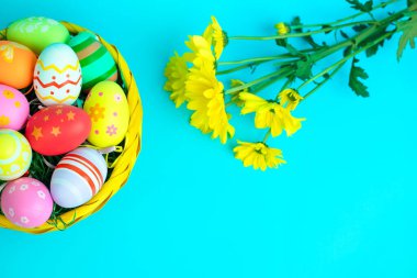 Canlı bir sepet renkli Paskalya yumurtasına parlak mavi arka planda taze sarı çiçekler eşlik ediyor. Neşeli ve neşeli bir bahar tatili kompozisyonu yenilenmeyi ve neşeyi simgeliyor..