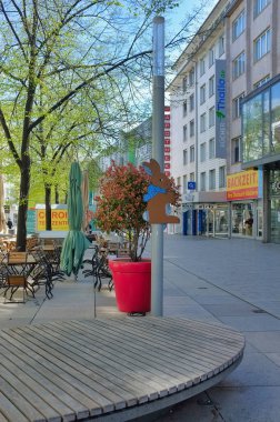 Thalia.de adında bir dükkan ve BACKZEIT Kafe 'nin yer aldığı bir sokak manzarası. Ağaçlar kaldırıma dizilir ve şemsiyeli açık hava masaları rahatlamaya davetiye çıkarır. Kırmızı bir saksı bir çalı tutar ve tahta bir tavşan süslemesi sahneye cazibe katar..