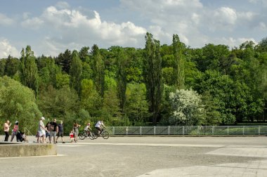 Görüntü, insanların eğlence amaçlı aktiviteler yaptığı canlı bir park ortamını gösteriyor. Bazıları bisiklete biniyor, bazıları yürüyor ve bazıları bebek arabalarını itiyor, hareketli ama sakin bir atmosfer yaratıyorlar. Sahne yemyeşil ağaçlar ve...