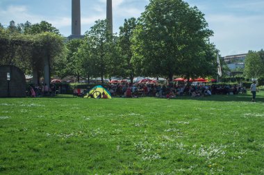 Fotoğraf, küçük beyaz çiçeklerle benekli uçsuz bucaksız yeşil çimlerle dolu canlı bir park yakalıyor. Arka planda, insanlar ağaçların ve kırmızı şemsiyelerin altında toplanarak hoş bir ortam yaratıyorlar. Çimlerin üzerinde renkli bir çadır ve iki kule