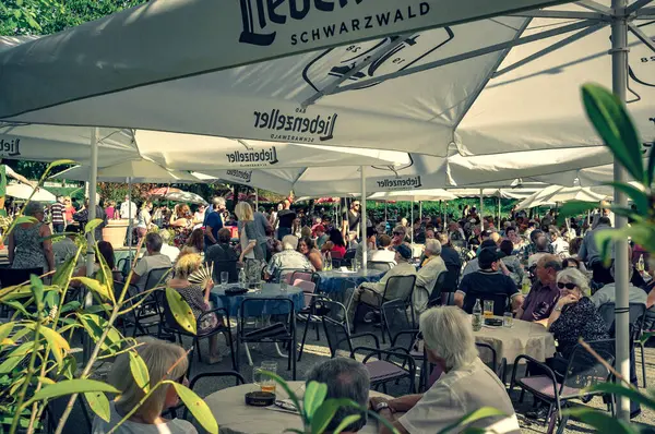 Görüntü, Lieberenzeller ile damgalanmış büyük beyaz şemsiyeler altında oturan müşterilerle dolu hareketli bir açık hava kafesi yakalıyor. İnsanlar içki içerken neşeli sohbetlere dalarlar. Mekan, hoş bir karşılama ortamı yayıyor.