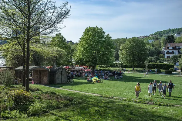 Görüntü, bir müzisyenin hem klavye hem de gitar çaldığı bir açık hava sahnesini tasvir ediyor. Ön planda çeşitli müzik aletleri ve ekipmanlar sergileniyor. Arka planda, büyük bir kalabalık beyaz şemsiyelerin altında toplanır, canlı olmanın tadını çıkarırlar.