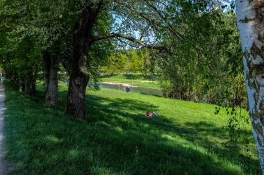 Fotoğrafta, güneşli bir günde çimenleri beyaz çiçeklerle süsleyen yemyeşil bir park sergileniyor. İnsanlar ağaçların ve kırmızı şemsiyelerin altında toplanırken renkli bir çadır canlılık katıyor. Uzakta, iki endüstriyel baca dinginlikle tezat oluşturuyor.