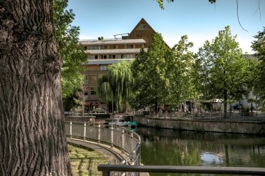 Fotoğraf, her iki taraftaki yemyeşil ağaçlarla çevrili sakin bir kanalı tasvir ediyor. Su sakindir, etrafındaki yaprakları yansıtır. Solda, arka planda parmaklıkları ve bir binası olan asfalt bir geçit var. Sahne sakin ve...