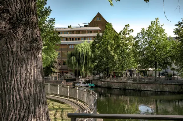 Fotoğraf, her iki taraftaki yemyeşil ağaçlarla çevrili sakin bir kanalı tasvir ediyor. Su sakindir, etrafındaki yaprakları yansıtır. Solda, arka planda parmaklıkları ve bir binası olan asfalt bir geçit var. Sahne sakin ve...