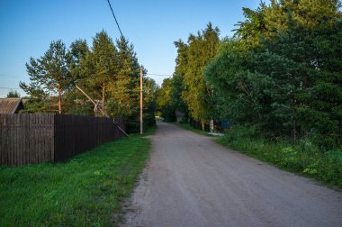 Bu görüntü, her iki tarafında ağaçlar ve yeşillikler olan kırsal toprak bir yol yakalıyor. Sol tarafta ahşap bir çit, sağ tarafta ise yemyeşil bitki örtüsü var. Yol mesafeye kadar uzanıyor, bir huzur ve yalnızlık hissi uyandırıyor.