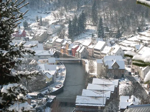 Almanya 'da sakin bir kış manzarası: karla kaplı ağaçlar, beyaz çatılı evler, karda derin lastik izleri ve parlak mavi-beyaz bir zemin. Güneş berrak gökyüzünde parlıyor, karlı kara parçasının üzerine huzur dolu bir parıltı saçıyor.