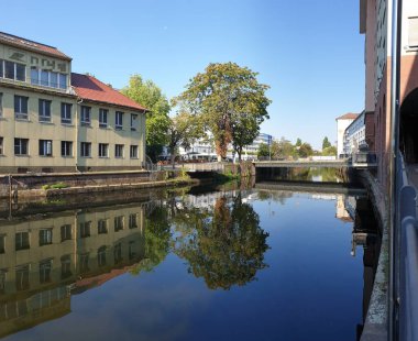 Binalarla çevrili bir nehir kırmızı çatılı ve kemerli bir yapıya sahip sarı bir evi yansıtır. Merkezdeki bir köprü ve ağaç manzarayı tamamlıyor..