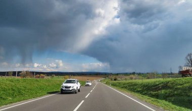 Bulutlu bir gökyüzünün altında çiçek açan ağaçlar ve yeşil tarlalarla dolu dolambaçlı bir yol. Yürüyüş ya da manzaralı sürüş için mükemmel bir yer..