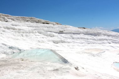 Büyüleyici bir doğa harikası, beyaz teraslar çağlayarak aşağı iniyor, berrak mavi gökyüzünün altında akan su ve mineral yataklarının nefes kesici bir sahnesini oluşturuyor.