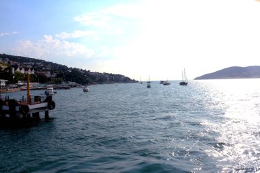 Ağaçlarla kaplı tepeleri olan sakin sularda bir rıhtım ve teknelerin yer aldığı sakin bir sahil manzarası Türkiye ve yukarıda parlak güneş ışığı