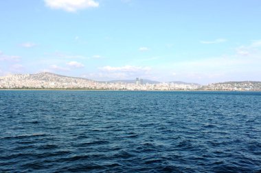 Görüntü, önünde engin mavi bir deniz bulunan sakin bir kıyı şehrini ve berrak bir gökyüzünün altındaki tepelik arazide yerleşik olan kentsel binaları gösteriyor.