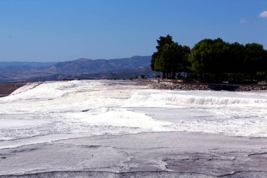 Büyüleyici bir doğa harikası, beyaz teraslar çağlayarak aşağı iniyor, berrak mavi gökyüzünün altında akan su ve mineral yataklarının nefes kesici bir sahnesini oluşturuyor.