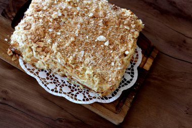 Nefis bir Napolyon keki, diğer adıyla mili-feuille, ahşap bir tahtanın üzerinde beyaz bir altlık üzerinde oturur, pullu tabakalarını, kremsi dolgusunu ve ufalanmış üstünü gösterir..