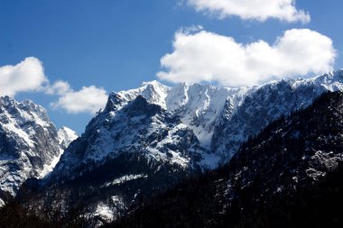 Görkemli karla kaplı dağlar parlak mavi bir gökyüzüne karşı yükselirler tüylü beyaz bulutlarla benekli, çarpıcı bir alp manzarası yaratırlar..
