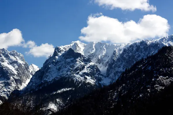 Görkemli karla kaplı dağlar parlak mavi bir gökyüzüne karşı yükselirler tüylü beyaz bulutlarla benekli, çarpıcı bir alp manzarası yaratırlar..