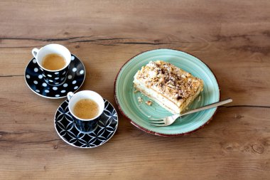 İki fincan espresso desenli fincanda ve çay tabağında yeşil bir tabakta çatallı bir pasta diliminin yanında oturuyor, hepsi de sıcak ahşap bir masada..