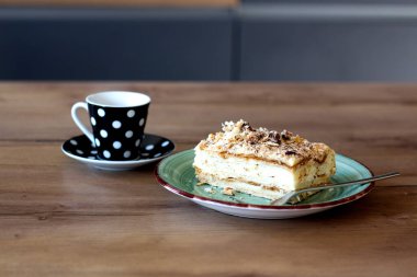 İki fincan espresso desenli fincanda ve çay tabağında yeşil bir tabakta çatallı bir pasta diliminin yanında oturuyor, hepsi de sıcak ahşap bir masada..