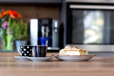 İki şık puantiyeli bardak, ahşap tezgahın üzerindeki lezzetli bir dilim pastanın yanında duruyor. Parlak bir mutfak ortamında, bulanık aletler ve çiçeklerle..