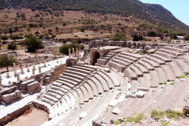 Bu resim, tepelerin ve seyrek bitki örtüsünün ortasındaki taş koltuklu ve sütunlu antik bir amfi tiyatroyu tasvir ediyor..
