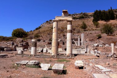 Görüntü, kuru bir yamaç ve bitki örtüsüyle çevrili parlak mavi bir gökyüzünün altında duran antik kalıntıları gösteriyor..