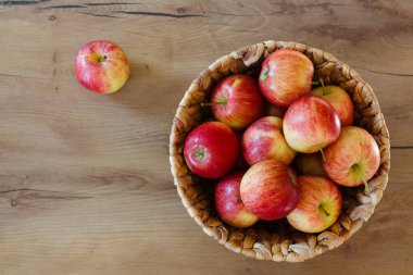 Bir dokuma sepet, ahşap bir yüzey üzerinde canlı kırmızı ve sarı elmalar taşar, bir başıboş elma dolu konteynır yanında dinlenir.