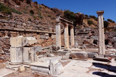 Fotoğrafta, tarihi bir arkeolojik alanı temsil eden, temiz bir gökyüzünün altında ayakta duran sütunları ve kırık taş yapıları bulunan antik kalıntılar sergileniyor..