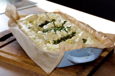 Erimiş mozzarella, canlı yeşil pesto kıvrımları ve taze biberiye filizleriyle kaplı dikdörtgen bir Focaccia, ahşap bir yüzey üzerinde mavi bir kapta oturur.