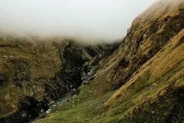 Derin bir dağ vadisi aşağıdaki çimenli ve kayalık arazide yolunu oyan kayalık bir dere boyunca tepeleri kaplayan alçak sislerle doludur.