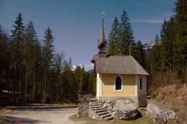 Siyah çatılı ve kulesi olan büyüleyici sarı şapel Avusturya Alplerinde, açık mavi bir gökyüzünün altında, ebedi yeşil ağaçların arasında huzur içinde duruyor.