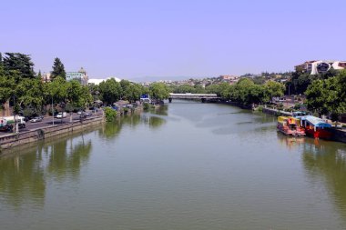 Geniş, sakin bir nehir, güneşli bir günde kıyılarını saran binalar, ağaçlar, tekneler ve bir köprü ile berrak bir gökyüzünün altındaki kentsel manzarayı yansıtır.