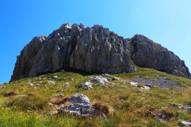 Pürüzlü uçurumları olan yüksek bir kayalık dağ, parlak mavi bir gökyüzünün altında çimenli bir yamacın üzerinde yükseliyor. Engebeli ve sakin bir manzara.