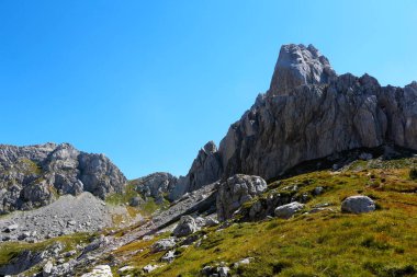 Görkemli dağlar manzaraya hükmediyor. Kayalık tepeleri, güneşli bir günde açık mavi bir gökyüzünün altındaki yeşil çimlerle çelişiyor.