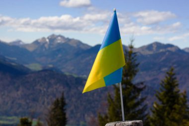 Parlak mavi ve sarı Ukrayna bayrağı, mavi gökyüzünün altında tüylü beyaz bulutlarla süslenmiş çarpıcı bir dağ manzarasına gururla karşı durmaktadır.