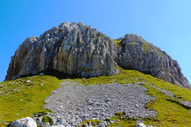 Karadağ 'daki açık mavi gökyüzüne karşı heybetli kayalık bir dağ yükseliyor. Yeşil yamaçları engebeli ve etkileyici bir zirveye çıkıyor.