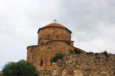 Kırmızı kiremitli kubbe ve haçla süslenmiş tarihi Jvari Manastırı yayılmış, bulutlu bir gökyüzünün altındaki tepede görkemli bir şekilde duruyor.