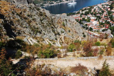 Yüksek bir açıyla, Karadağ 'da engebeli bir dağa tırmanan bir yılan yolu, büyük bir yolcu gemisi ve aşağıda yuva yapmış büyüleyici bir sahil kasabasının manzarasını sunuyor.