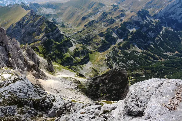 Nefes kesen bir manzara, keskin, kayalık oluşumlar ve dağınık bir ışık altında yayılmış yeşilliklerle karakterize edilmiş sert ve görkemli bir dağ manzarasını gözler önüne serer.