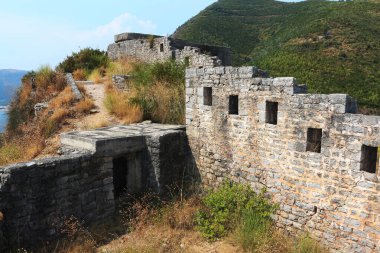 Taş kale, deniz manzaralı bir uçurum üzerinde, arkasında yemyeşil bir yamaç bulunan bir tüneği harap eder.