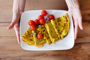 İki el katlanmış sarı krepli beyaz bir tabağı tutuyor, filizli kiraz domatesleri ve kekik ile süslenmiş, ahşap bir yüzey üzerinde.