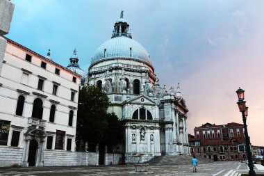Büyük, beyaz Bazilika di Santa Maria della Salute esmer bir gökyüzü altında ikonik kubbeleri ve Venedik mimarisinin karmaşık bir yüzü altında etkili bir şekilde duruyor. Tarihsel binalar önplandaki açık asfaltlı alanı çerçeveliyorlar