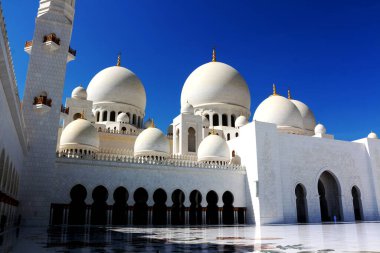 Şeyh Zayed Büyük Camii 'nin ihtişamı, el değmemiş beyaz mermer cephesi, zarif kubbeleri ve çarpıcı İslami tasarım unsurlarıyla canlı mavi bir zemin oluşturmaktadır. Avlu zemininde yansımalar.
