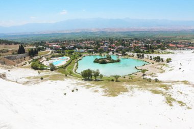 Pamukkale 'nin saf beyaz travertin teraslarına karşı parlak turkuaz havuzların çarpıcı kontrastı. Yakınlardaki bir kasaba ve sisli bir dağ sırası engin, berrak bir mavi gökyüzünün altında oturur..