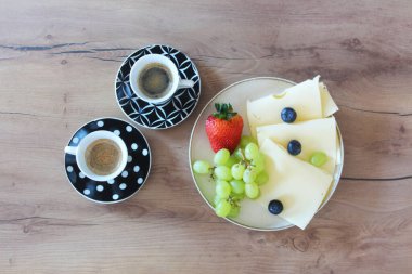 Bir tabak peynir, tatlı üzüm, canlı bir çilek ve koyu yaban mersininin yanında iki fincan zengin espresso bulunan ahşap bir masada basit bir kahvaltı veya atıştırmalık düzenlenir.