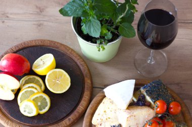 İki tahta tahta tahta, canlı kırmızı bir elma sulu limon dilimleri, çeşitli peynirler ve parlak vişneli domateslerle dolu. Bir saksı yeşil bitki ve bir bardak zengin kırmızı şarap kırsal bir ahşap masada sahnenin cazibesini artırır.