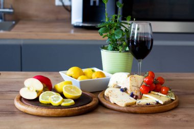 Kırsal ahşap bir masada iki akasya ekmek tahtası bulunur. Birinde dilimlenmiş elma ve limon sergilenirken, diğerinde peynir ve vişneli domates bulunuyor. Bir bardak kırmızı şarap ve saksı bitkisi bu davetkar natürmortu tamamlıyor.