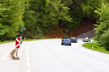 Avusturya 'da aydınlık bir günde yeşil yapraklarla çevrili kıvrımlı bir yolda üç araba dolaşır.