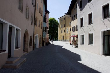 Rattenberg 'deki huzurlu bir kaldırım taşı caddesi büyüleyici, renkli binalarla çerçevelenmiş ve çeşitli mimari detaylara sahip. Gölgeler, parlak bir gökyüzünün altındaki kaldırım boyunca uzanır, Avrupa 'nın bu tuhaf manzarasında, gözleri uzak bir ağaca götürür.